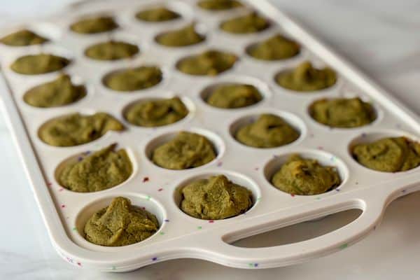 Angled view of green plant-based baby-led weaning mini muffins freshly baked in a white confetti mini muffin tray on a marble surface.