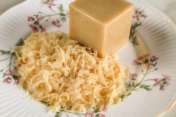 A block of homemade vegan parmesan cheese alongside a generous pile of freshly grated vegan parmesan, displayed on a vintage white ribbed ceramic plate with pink floral details and gold trim on a light background.