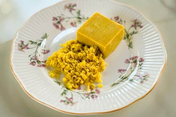 A block of homemade vegan cheddar cheese alongside a pile of freshly grated vegan cheddar, displayed on a vintage white floral plate with gold trim on a marble surface.