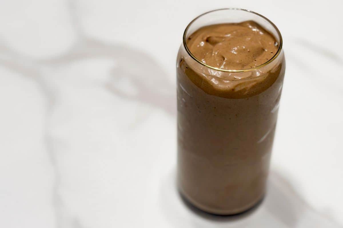Overhead view of a thick and creamy high protein plant-based chocolate avocado smoothie in a gold rimmed glass can cup on a white marble surface with dramatic shadow lighting.