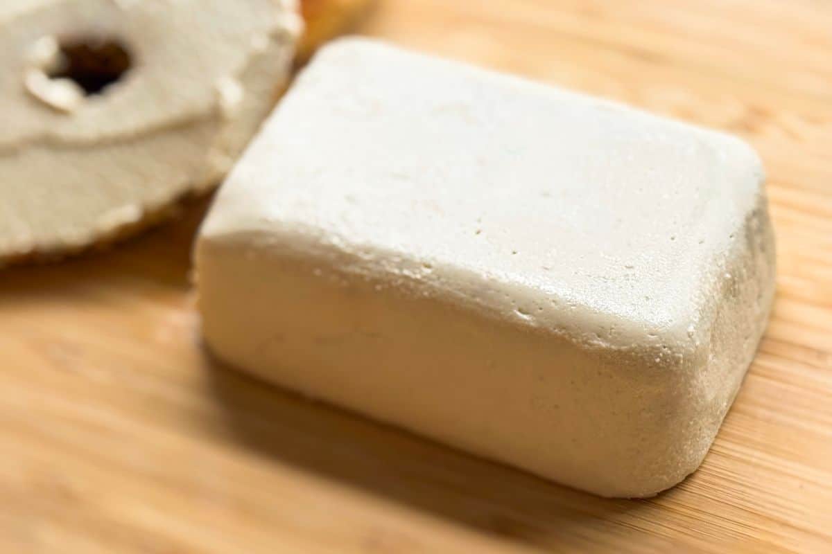 Angled close up of a homemade plant-based cream cheese block on a bamboo cutting board with a sliced toasted bagel with cream cheese spread on it in a blurred in the background.