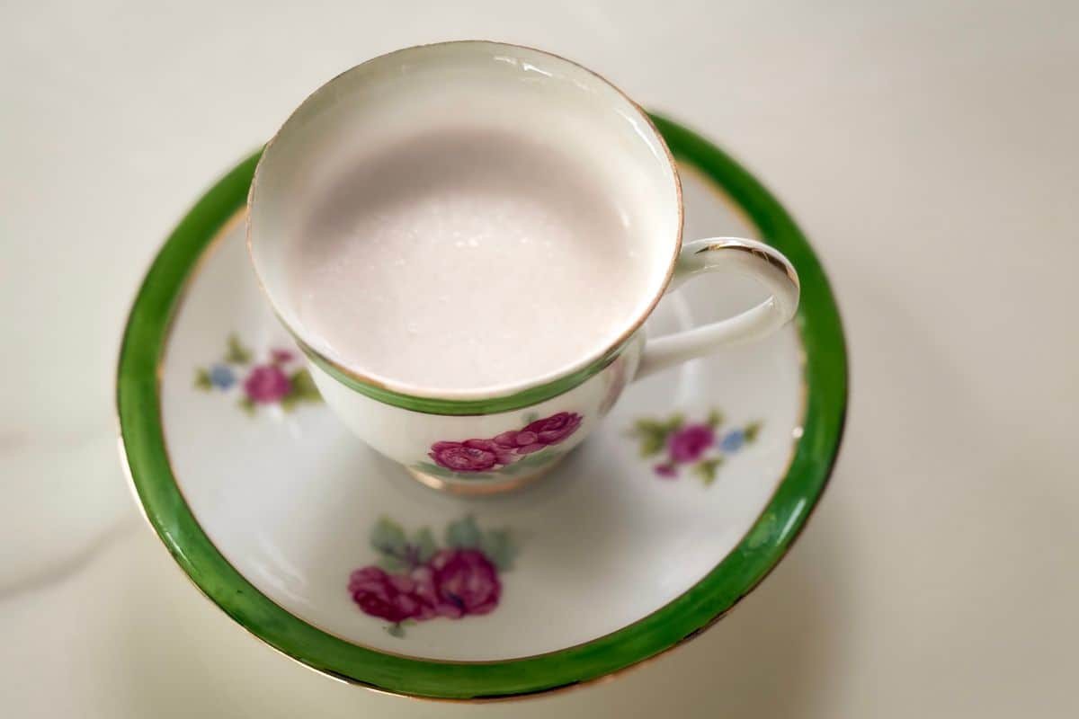 A smooth, pale vegan cream substitute served in a vintage white and green floral teacup with pink rose details and gold trim, resting on a matching saucer against a soft white background, photographed from above.