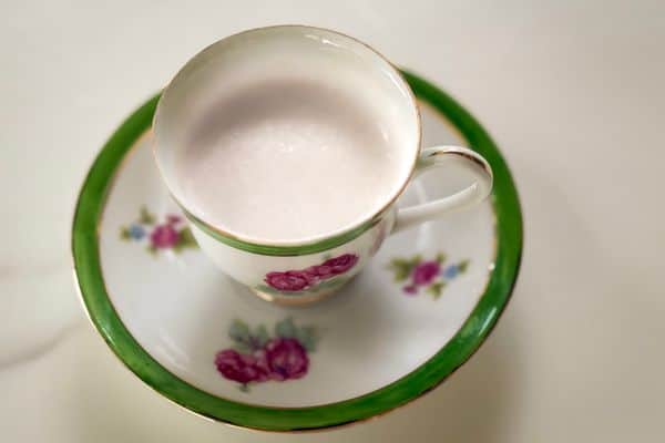 A smooth, pale vegan cream substitute served in a vintage white and green floral teacup with pink rose details and gold trim, resting on a matching saucer against a soft white background, photographed from above.