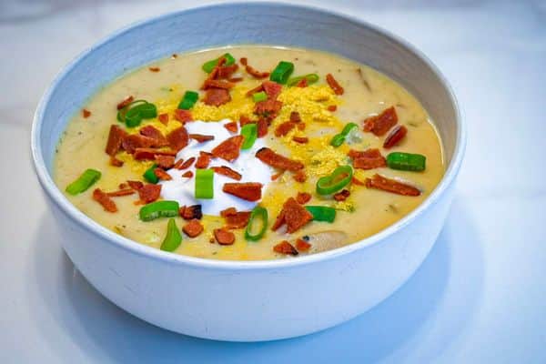 A creamy vegan loaded baked potato soup served in a rustic white ceramic bowl, topped with plant-based bacon bits, sliced green onions, a dollop of vegan sour cream, and a sprinkle of nutritional yeast, photographed on a marble surface.