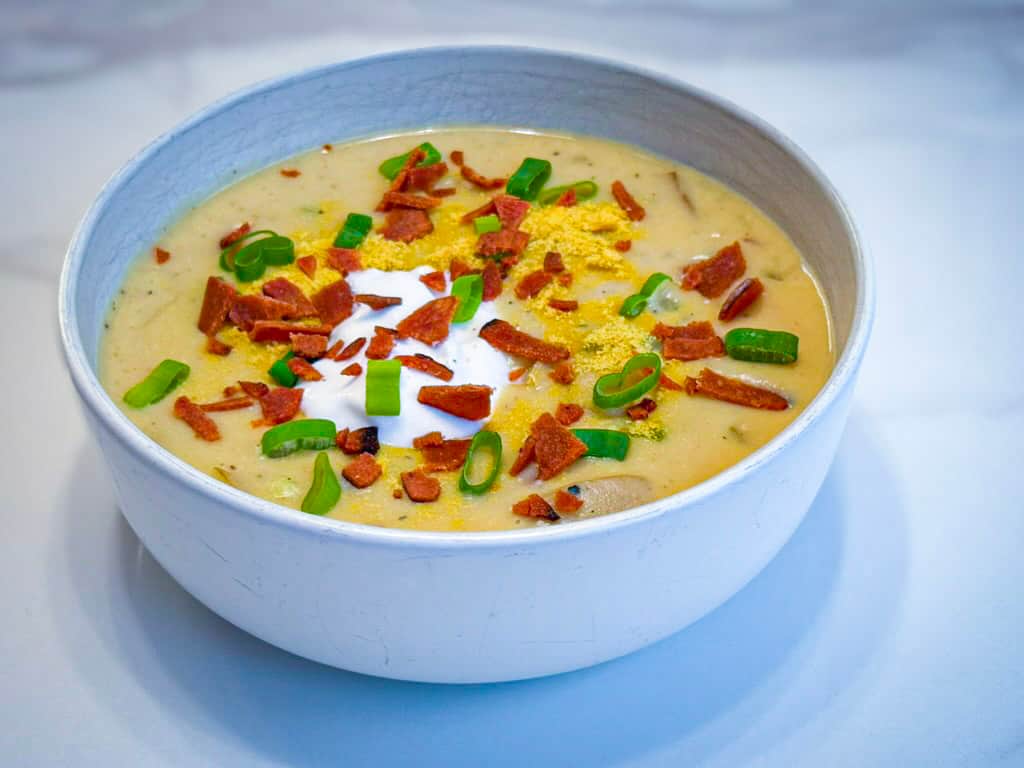A creamy vegan loaded baked potato soup served in a rustic white ceramic bowl, topped with plant-based bacon bits, sliced green onions, a dollop of vegan sour cream, and a sprinkle of nutritional yeast, photographed on a marble surface.