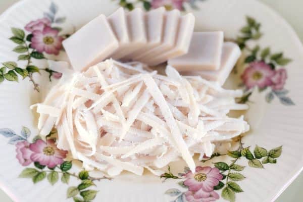 A block of homemade vegan mozzarella cheese sliced into pieces alongside a pile of freshly shredded vegan mozzarella, displayed on a vintage white floral plate with pink rose and green leaf details on a light grey surface.