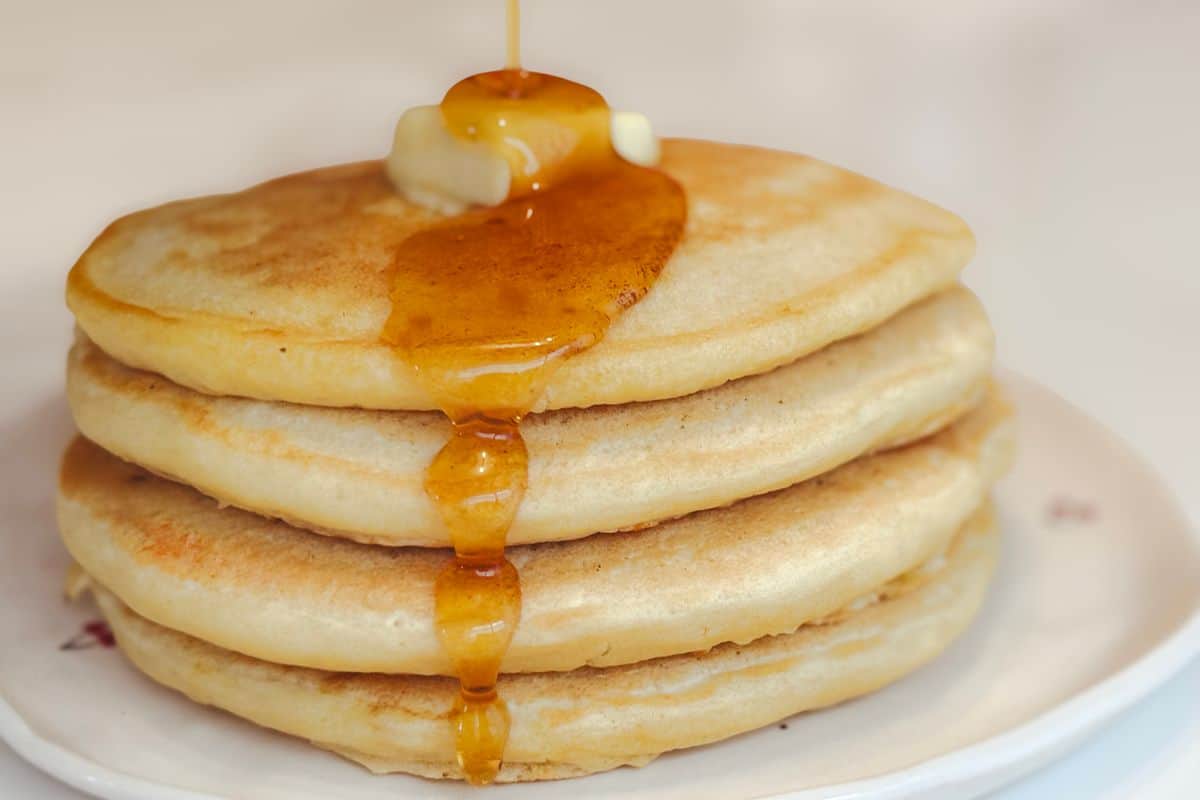 Maple syrup being poured over a stack of four fluffy vegan buttermilk pancakes topped with a pat of plant-based butter on a white cherry patterned plate.