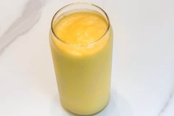 Overhead view of a creamy high protein plant-based passion fruit and mango smoothie in a gold rimmed glass can cup on a white marble surface, vibrant yellow and orange in color.