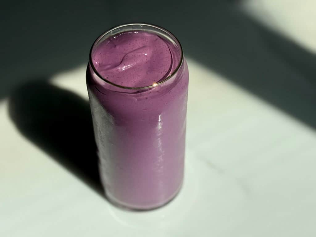 Overhead view of a creamy high protein plant-based peanut butter and jelly smoothie in a glass can cup on a dark marble surface with dramatic shadow lighting.