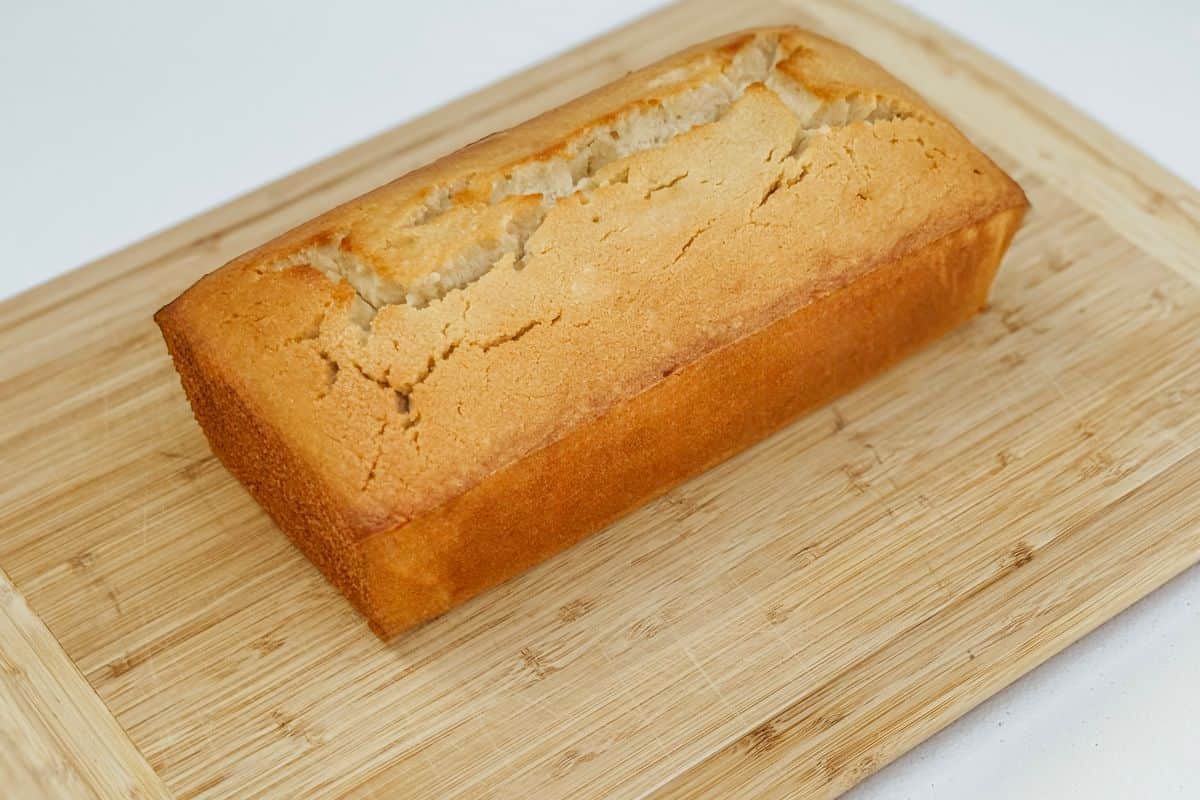 A golden-brown vegan pound cake with a perfectly cracked top crust, resting on a natural bamboo cutting board against a clean white background.