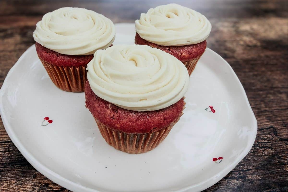 Three fluffy vegan red velvet cupcakes topped with swirled dairy-free cream cheese frosting on a white cherry patterned plate on a rustic wooden surface.