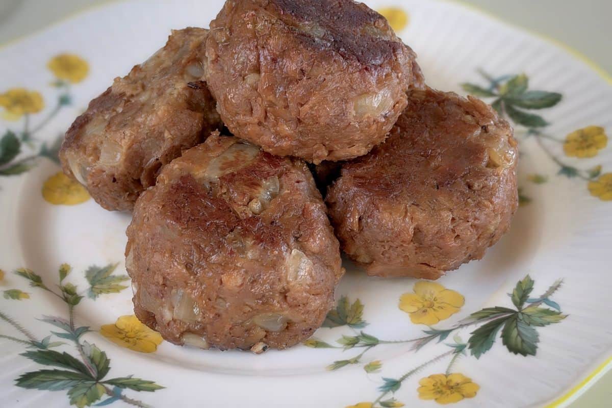 A pile of golden-brown pan-seared vegan meatballs with a hearty, textured exterior and visible onion pieces, stacked on a vintage white ceramic plate with a yellow floral botanical pattern on a light background.