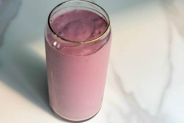 Overhead view of a creamy high protein plant-based strawberry banana smoothie in a glass can cup with a gold rim on a white marble surface.
