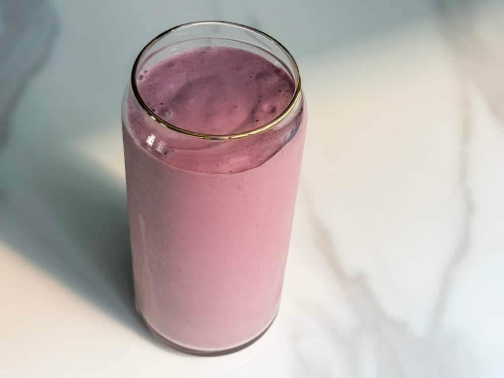 Overhead view of a creamy high protein plant-based strawberry banana smoothie in a glass can cup with a gold rim on a white marble surface.