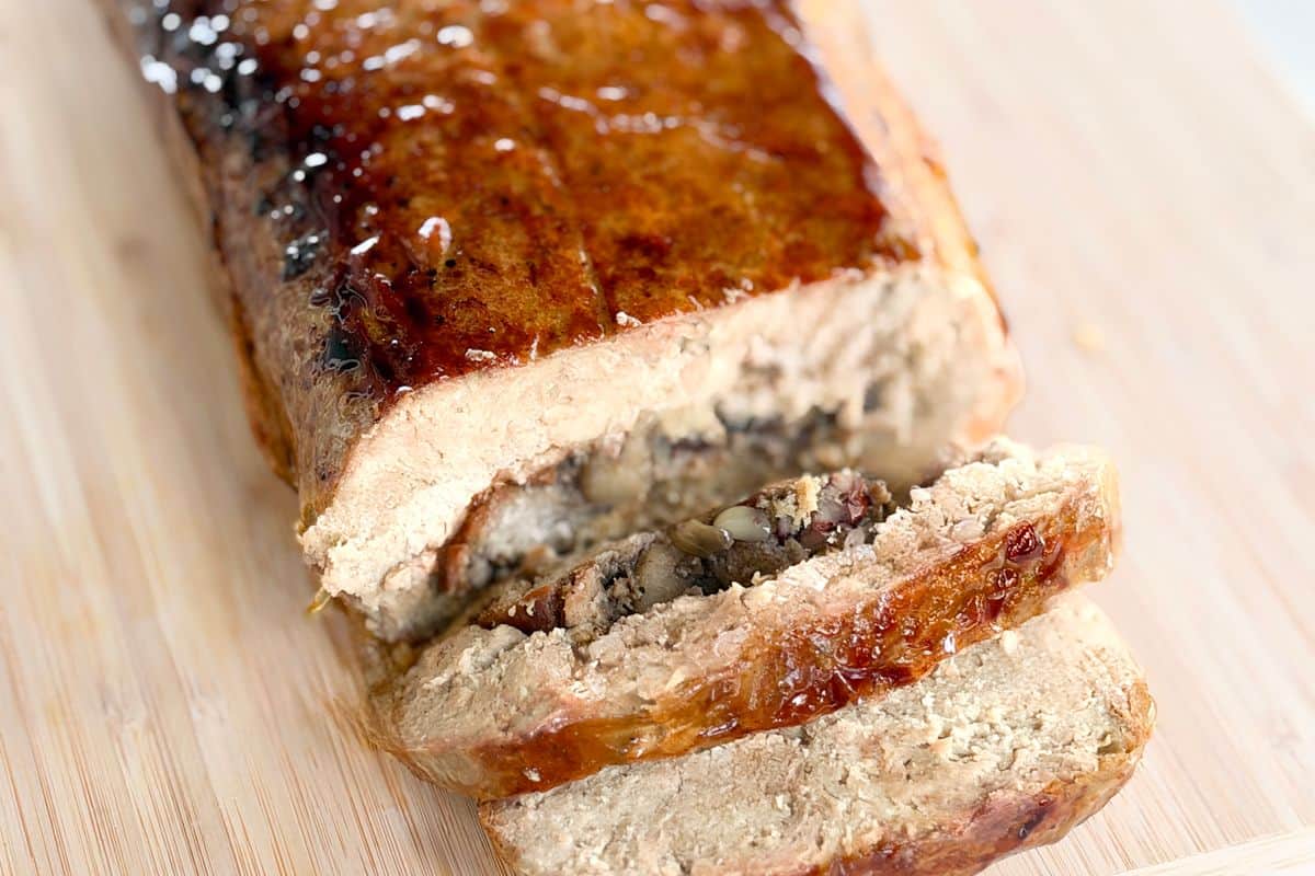 Sliced stuffed vegan roast with a glossy maple-soy glaze on a bamboo cutting board, showing the mushroom and pecan stuffing spiraled through the center.