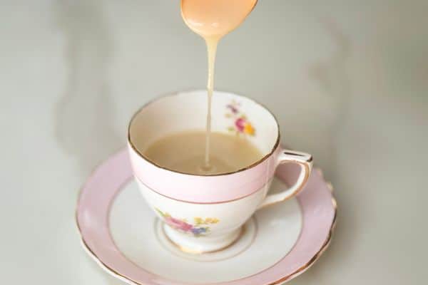 A golden stream of homemade vegan sweetened condensed milk being poured from a spoon into a vintage pink and white floral teacup with gold trim, resting on a matching saucer against a soft grey background.