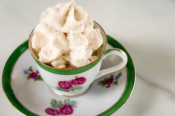 A generous swirl of homemade vegan whipped cream piled high in a vintage white and green floral teacup with gold trim, resting on a matching saucer against a soft grey background.
