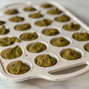 Angled view of green plant-based baby-led weaning mini muffins freshly baked in a white confetti mini muffin tray on a marble surface.