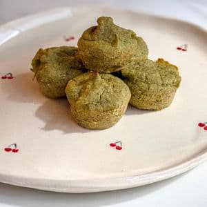 A pile of small green plant-based baby-led weaning mini muffins stacked on a white cherry patterned plate, soft and perfectly sized for baby fingers.