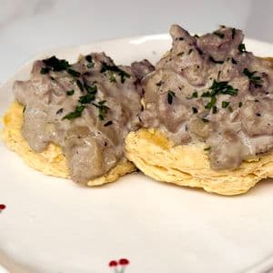 Two flaky vegan buttermilk biscuits smothered in creamy plant-based beyond meat gravy garnished with fresh chopped parsley on a white cherry patterned plate on a marble surface.