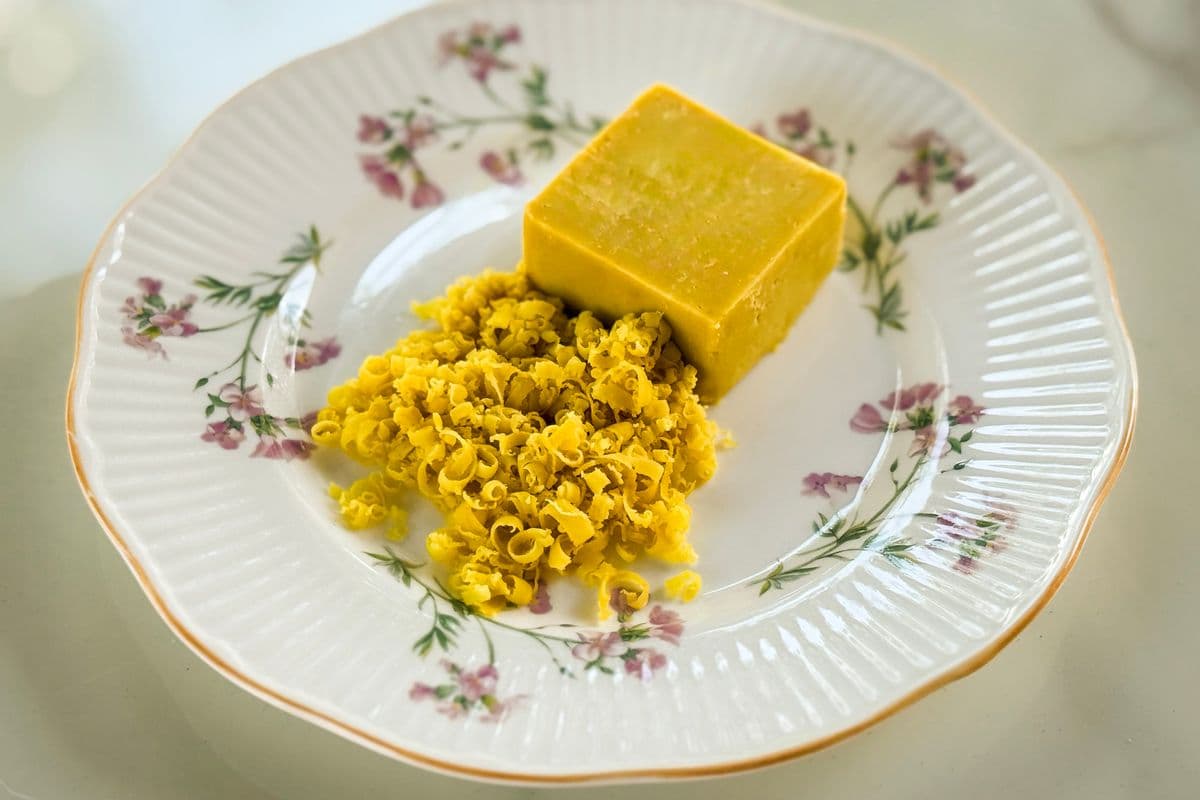 A block of homemade vegan cheddar cheese alongside a pile of freshly grated vegan cheddar, displayed on a vintage white floral plate with gold trim on a marble surface.