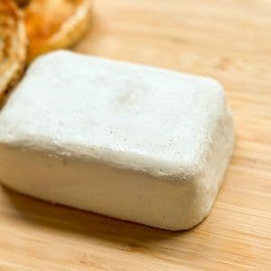 Close up front view of a homemade plant-based cream cheese block on a bamboo cutting board with a toasted bagel visible in the background.
