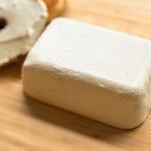 Angled close up of a homemade plant-based cream cheese block on a bamboo cutting board with a sliced toasted bagel with cream cheese spread on it in a blurred in the background.