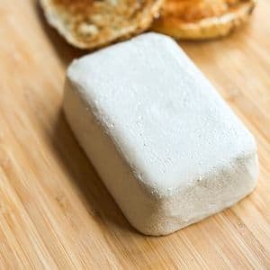 A block of homemade plant-based cream cheese on a bamboo cutting board with a toasted bagel blurred in the background.