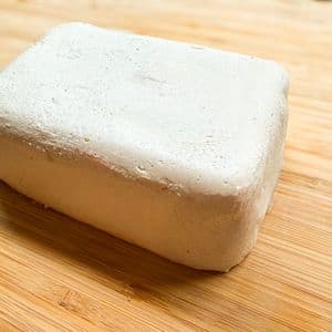 A block of homemade plant-based cream cheese on a bamboo cutting board, showing a smooth white exterior with a slightly textured surface.