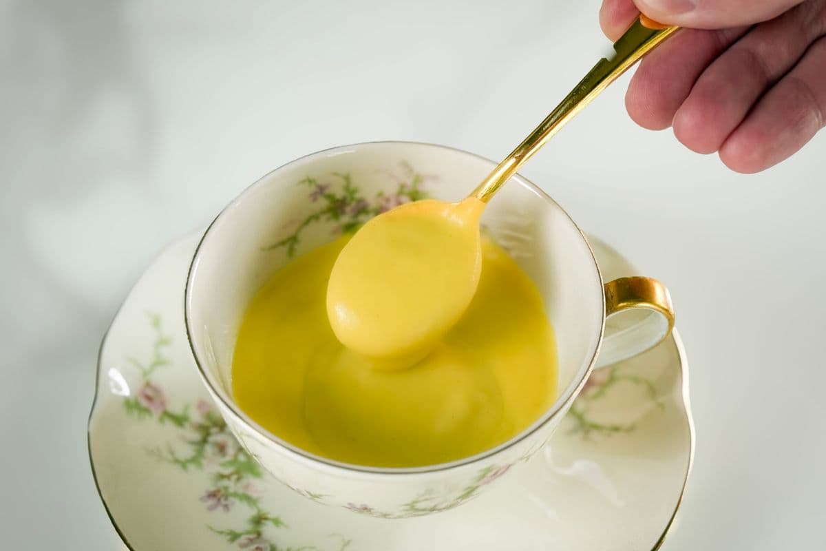 A hand holding a gold spoon lifting a scoop of rich, golden vegan hollandaise sauce from a vintage white floral teacup with gold trim on a matching saucer, photographed against a soft white background.