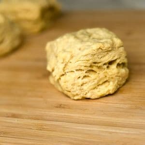 Close up of a single fluffy vegan biscuit on a bamboo cutting board showing its flaky layered texture with another biscuit blurred in the background.