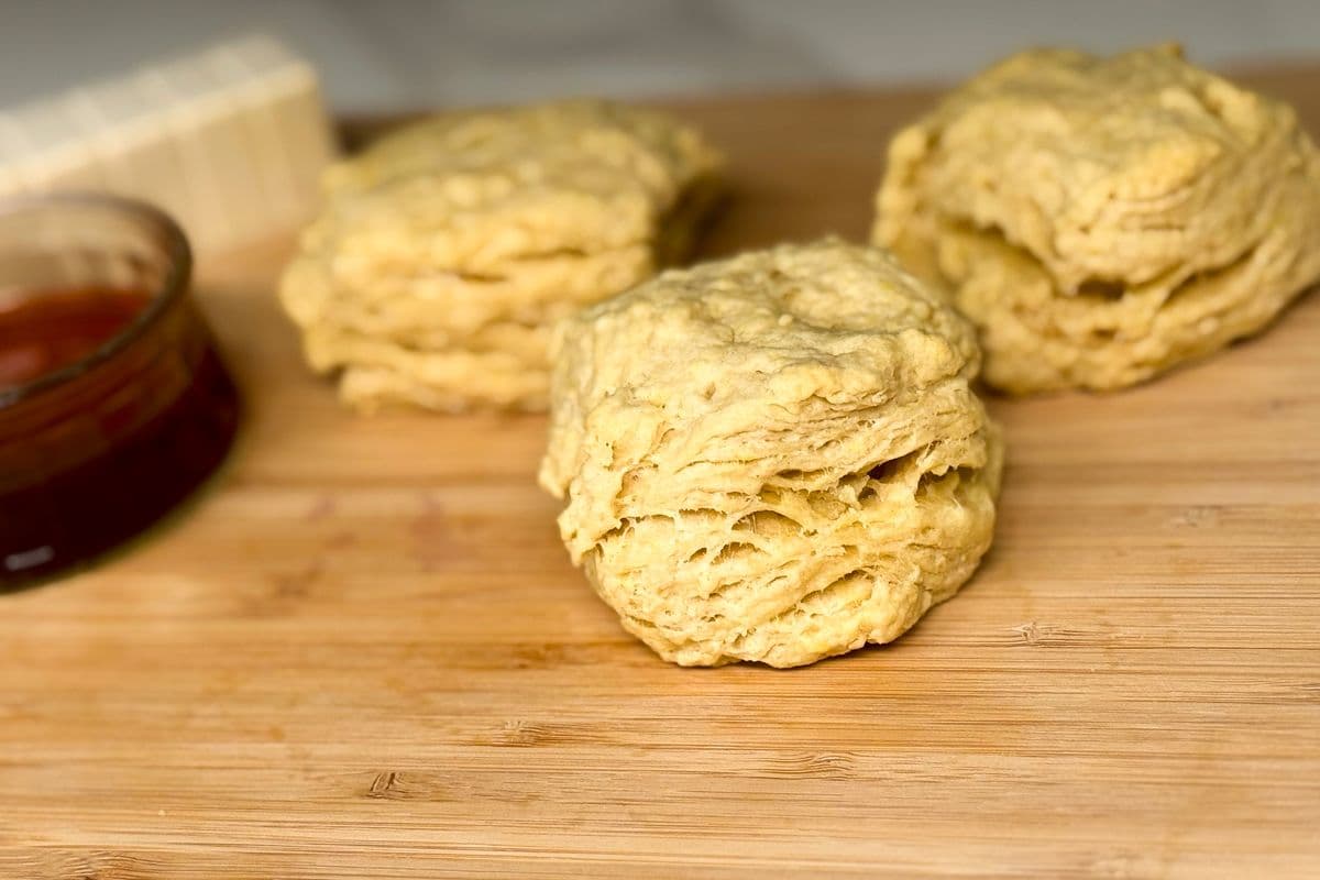Three fluffy layered vegan biscuits on a bamboo cutting board with a small bowl of maple syrup and a block of plant-based butter blurred in the background.