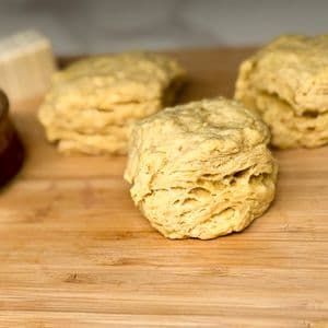 Three fluffy layered vegan biscuits on a bamboo cutting board with a small bowl of maple syrup and a block of plant-based butter blurred in the background.