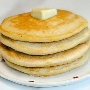 A stack of four fluffy golden vegan buttermilk pancakes topped with a pat of homemade plant-based butter on a white cherry patterned plate on a white surface.