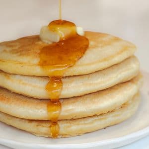Maple syrup being poured over a stack of four fluffy vegan buttermilk pancakes topped with a pat of plant-based butter on a white cherry patterned plate.