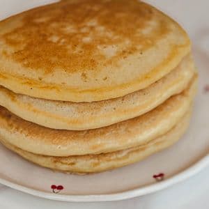 A stack of four golden fluffy vegan buttermilk pancakes on a white cherry patterned plate showing the perfectly golden brown tops and fluffy layers.