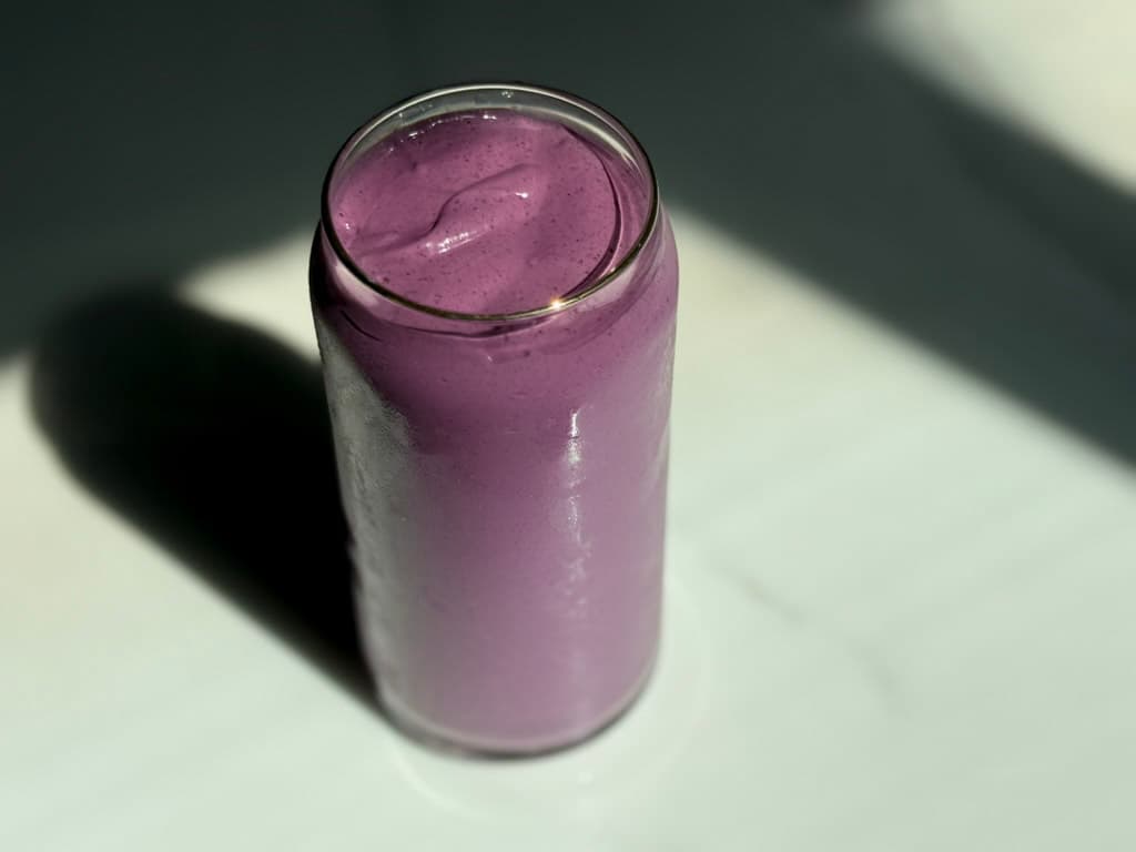 Overhead view of a creamy high protein plant-based peanut butter and jelly smoothie in a glass can cup on a dark marble surface with dramatic shadow lighting.