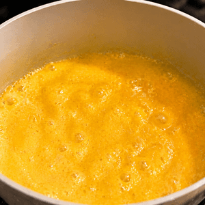 Overhead view of golden vegan egg yolk sauce bubbling and simmering in a white saucepan on a gas stovetop.