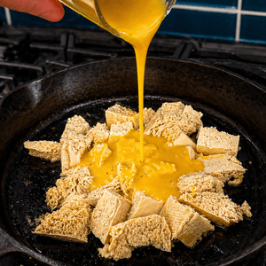 Golden vegan egg yolk sauce being poured over chunks of firm tofu in a cast iron skillet on a gas stovetop, in the process of making a plant-based tofu scramble.