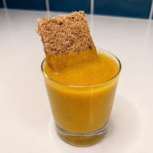 A glass of golden vegan egg yolk sauce with a piece of multigrain toast dipped into it, served on a white surface with blue tile background.