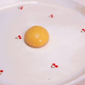 A single golden plant-based egg yolk sphere sitting on a white cherry patterned plate, created using spherification with Calcium Lactate and a Sodium Alginate bath.