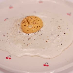 A plant-based fried egg on a white cherry patterned plate, featuring a golden vegan egg yolk sphere centered on a round animal-free egg white, seasoned with cracked black pepper.