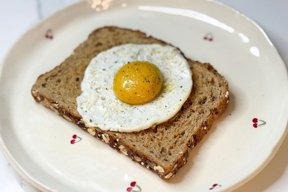 Plant-based fried egg on multigrain toast served on a white cherry plate, featuring a golden vegan egg yolk sauce on a vegan egg white, seasoned with black pepper.