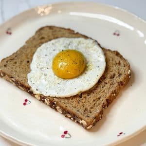 Plant-based fried egg on multigrain toast served on a white cherry plate, featuring a golden vegan egg yolk sauce on a vegan egg white, seasoned with black pepper.