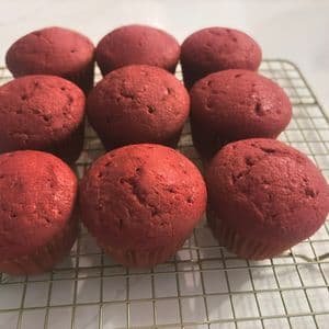 Nine deep red fluffy vegan red velvet cupcakes cooling on a gold wire cooling rack on a white marble surface, showing their perfectly domed tops before frosting.
