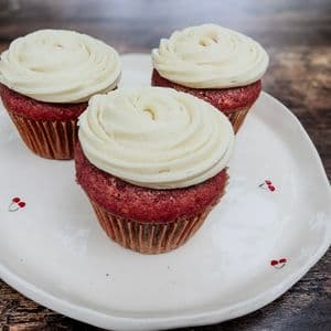 Three fluffy vegan red velvet cupcakes topped with swirled dairy-free cream cheese frosting on a white cherry patterned plate on a rustic wooden surface.