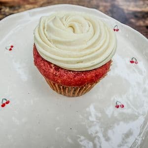 A single fluffy vegan red velvet cupcake topped with a swirled rosette of dairy-free cream cheese frosting on a white cherry patterned plate on a rustic wooden surface.