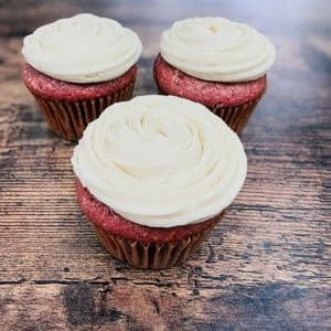 Three fluffy vegan red velvet cupcakes topped with swirled dairy-free cream cheese frosting in brown cupcake liners on a rustic wooden surface.