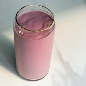 Overhead view of a creamy high protein plant-based strawberry banana smoothie in a glass can cup with a gold rim on a white marble surface.