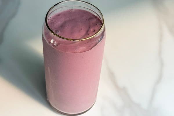 Overhead view of a creamy high protein plant-based strawberry banana smoothie in a glass can cup with a gold rim on a white marble surface.